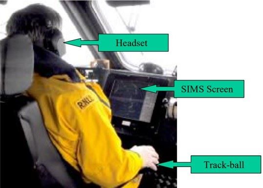 A photo of an RNLI crewman wearing a headset and a yellow jacket in a lifeboat. The photo is taken from behind, looking over the right shoulder and showing the position of the computer screen and the tracker ball for operating it.